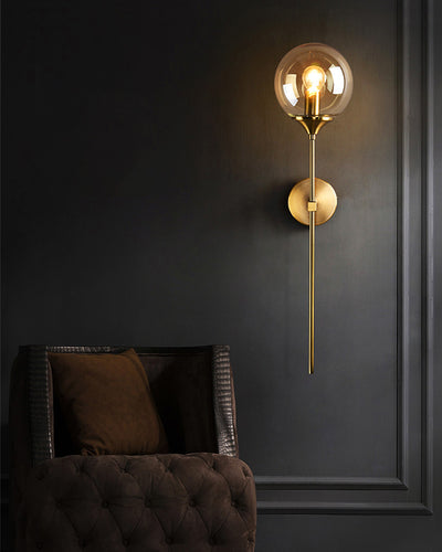 A nordic wall lamp with a long golden rod on a gray wall next to a brown couch