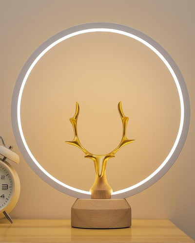 A light lamp with a golden deer, wooden base and a halo light on a night stand next to an alarm clock